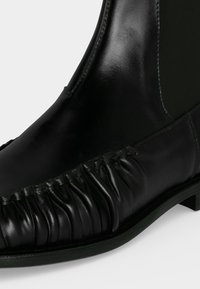 Gros plan d'une botte cheville en cuir noir avec des détails froncés sur le côté et un empiècement élastique pour un enfilage facile.