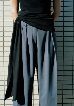 Persoon draagt losse grijze geplooide broek en een zwart topje met een lange zwarte stoffen doek, staand tegen een betegelde muur.