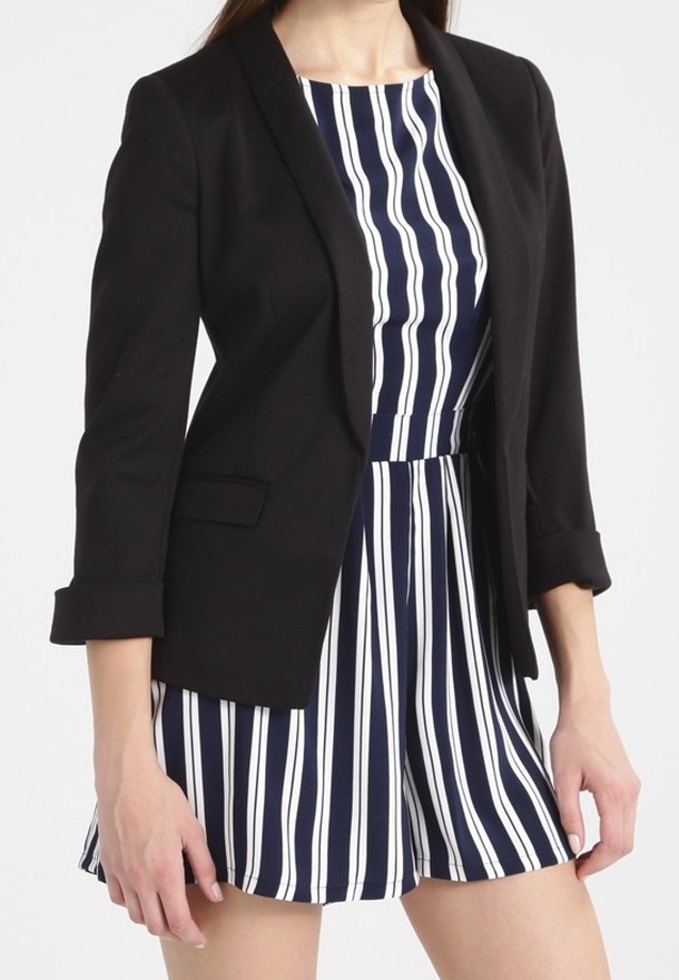 Femme portant un blazer noir par-dessus une combinaison sans manches à rayures verticales bleu marine et blanc, debout devant un fond uni.