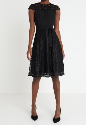 Femme portant une robe en dentelle noire mi-longue avec des manches courtes, une ceinture en tissu nouée à la taille et des talons pointus noirs.