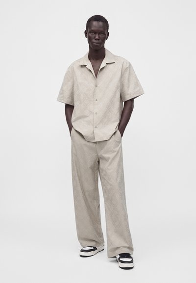 Homme portant une chemise beige à manches courtes à motifs et un pantalon ample assorti avec des baskets noires et blanches, debout devant un fond blanc.
