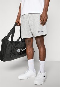 Grey cotton shorts with an embroidered "Champion" logo, paired with white sneakers and black duffel bag. Model holds bag with one hand.