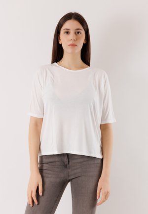 Giovane donna con capelli castani lunghi, che indossa una camicia bianca a maniche corte e jeans grigi, in piedi contro uno sfondo chiaro e uniforme.