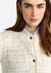 Giacca in tweed bianco con accenti dorati, tessuto testurizzato e tasca frontale. Presenta un colletto rotondo e una chiusura con bottone nero.