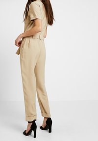 Combinaison beige à manches courtes, avec une ceinture à la taille et des jambes fuselées. Présente une texture lisse et des revers aux manches. Portée avec des talons noirs.