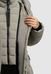 Jachetă puffer de culoare gri deschis, cu secțiuni matlasate, două buzunare cu fermoar, fermoar negru și o glugă mare, evidențiind o textură netedă și moale.