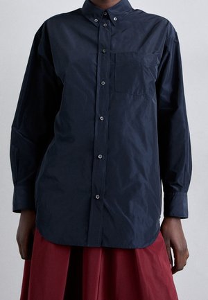 Person wearing a long-sleeve dark navy button-up shirt with a chest pocket and a red skirt, standing against a plain background.