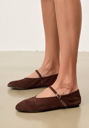 Meztelen lábak barna velúr Mary Jane stílusú lapos cipőben, kis arany csattal, sima bézs padlón.