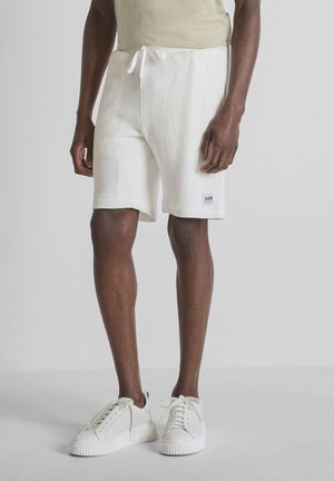 Hombre con pantalones cortos blancos texturizados con cordón y zapatillas blancas, posando contra un fondo gris liso.