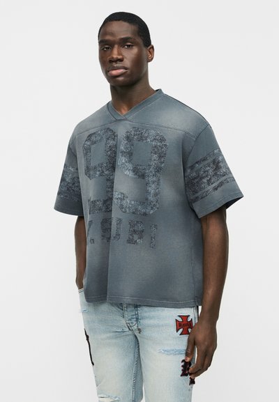 Νεαρός άνδρας που φοράει φθαρμένο γκρι φαρδύ πουκάμισο με το «99» και κείμενο στο μπροστινό μέρος, συνδυασμένο με ανοιχτόχρωμα σκισμένα τζιν με κόκκινες επιδιορθώσεις.