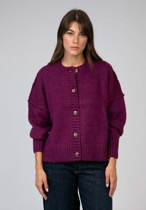 Femme portant un cardigan en tricot boutonné violet foncé avec des poignets et un ourlet côtelés, associé à un jean bleu foncé, se tenant contre un fond uni.