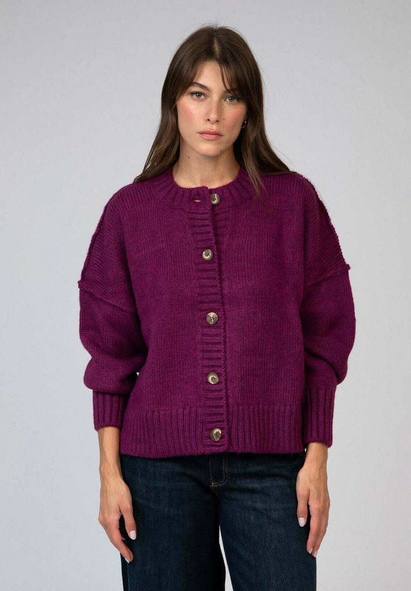 Femme portant un cardigan en tricot boutonné violet foncé avec des poignets et un ourlet côtelés, associé à un jean bleu foncé, se tenant contre un fond uni.