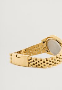 Χρυσό μεταλλικό ρολόι χειρός με συνδεδεμένο λουρί, που διαθέτει στρογγυλή κάσα και μηχανισμό κλεισίματος. Η πίσω όψη έχει λεία επιφάνεια με σήμανση.