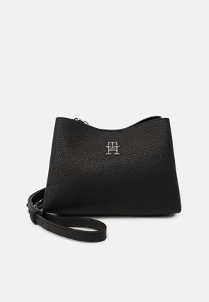 Petit sac bracelet en cuir texturé noir avec logo argenté "TH" et bandoulière amovible sur un fond clair uni.