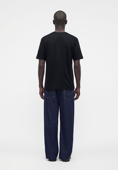 Homme debout, dos à la caméra, portant un t-shirt noir uni et un jean bleu foncé ample sur un fond blanc uni.