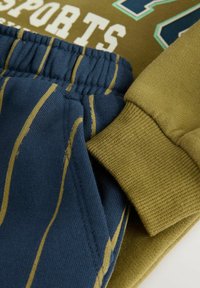 Olive green sweatshirt z wyraźnym napisem "SPORTS" oraz niebieskie spodnie z złotymi pionowymi paskami. Teksturowane i żebrowane mankiety na obu elementach odzieży.