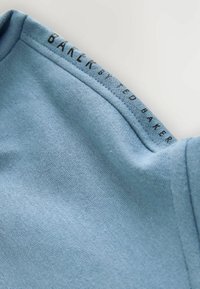 Szczegół niebieskiej bawełnianej koszulki, charakteryzującej się gładką strukturą i zaokrąglonym dekoltem. Nazwa marki „Baker by Ted Baker” nadrukowana na kołnierzu.