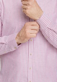 Camisa de botones con finas rayas rosas y blancas, con una textura suave y cierres de botón, mostrada sobre manos parcialmente entrelazadas.