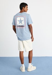 T-shirt en coton bleu clair avec une grande étoile blanche et "CONVERSE" imprimé en noir au dos. Associé à un short blanc et des baskets claires.