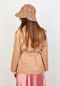 Impermeable beige con cinturón en la cintura y bolsillos laterales, combinado con un sombrero tipo bucket de cuadros en tonos camel, marrón y negro. El cabello del modelo cae liso.