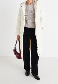 Long white puffer coat with a hood, worn over a textured beige sweater and black corduroy pants. Holding a small burgundy bag.