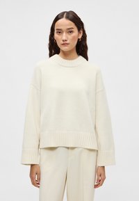 Krém színű, túlméretezett pulóver bordázott nyakkivágással és mandzsettával, puha anyagból készült. Világos színű nadrággal párosítva.