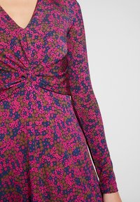 Sukienka z długim rękawem, z głębokim dekoltem w serek, zebranym detalem w pasie oraz kwiecistym wzorem w odcieniach różu, purpury i zieleni na ciemnym tle.