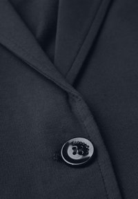 Námořnický blazer z látky s hladkým povrchem, s kulatým černým knoflíkem s vyraženým názvem značky. Ostré klopy a přizpůsobený střih.
