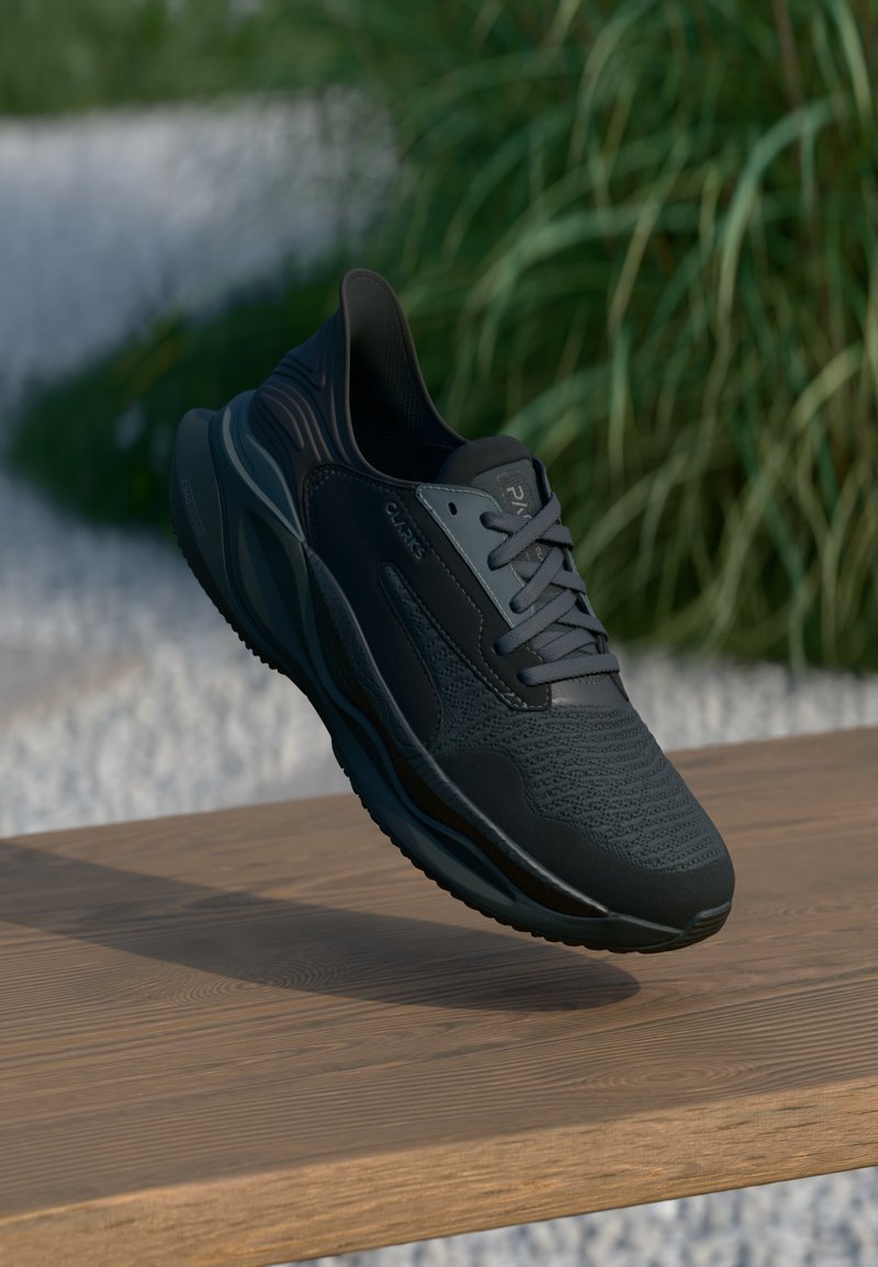 Baskets de sport noires avec une tige texturée et un design à lacets, présentées sur une surface en bois en extérieur avec de la verdure en arrière-plan.