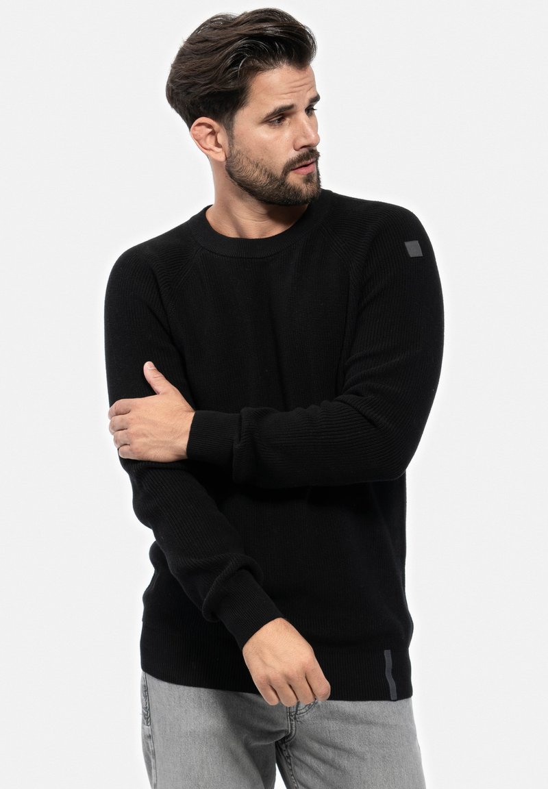 Schwarzer Rippstrickpullover mit langen Ärmeln, Rundhalsausschnitt und subtiler grauer Akzentuierung am Saum. Mann posiert mit einem Arm über dem anderen.