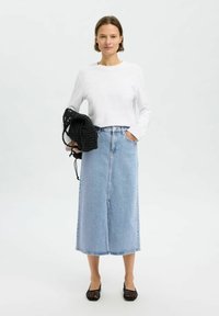 Blusa ligera blanca de manga larga combinada con una falda de mezclilla de talle alto y hasta la rodilla. La modelo lleva un bolso negro de mimbre y usa zapatos planos negros.