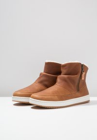 Botas de cuero marrón hasta el tobillo con cuello de piel blanca, cremallera lateral y superficie texturizada. Suela de goma blanca con un patrón de agarre duradero.