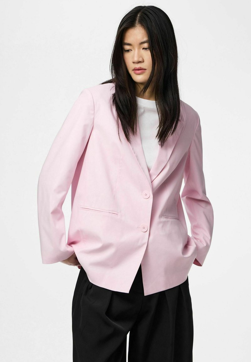 Femme asiatique aux cheveux noirs et lisses portant un blazer rose clair par-dessus un haut blanc et un pantalon noir, mains dans les poches, regardant vers le bas.