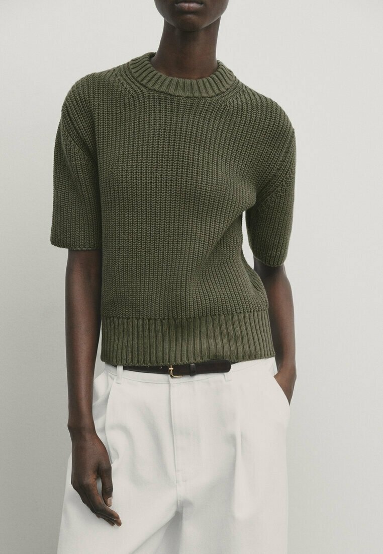 Olivengrøn kortærmet strikket sweater med struktureret mønster og ribkant, båret med hvide bukser og et sort bælte.
