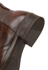 Botín de cuero marrón hasta el tobillo con punta redondeada, costuras detalladas y un tacón bajo apilado. La suela cuenta con goma texturizada para mayor agarre.