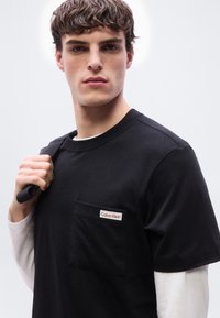 Νεαρός με κοντά, σγουρά καστανά μαλλιά φορώντας μαύρο T-shirt Calvin Klein πάνω από λευκή μακρυμάνικη μπλούζα, κρατώντας μια μαύρη τσάντα στον ώμο του.