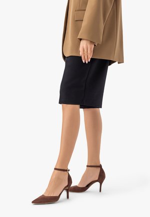 Kobieta w brązowych zamszowych szpilkach z paskiem na kostce, czarnej spódnicy do kolan i karmelowej marynarce, stojąca na jednolitym tle.