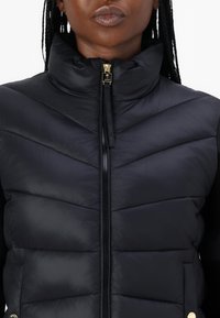 Vesta puffer neagră cu guler înalt, având un model diagonal matlasat, fermoar de culoare aurie și detaliu cu logo în partea stângă.