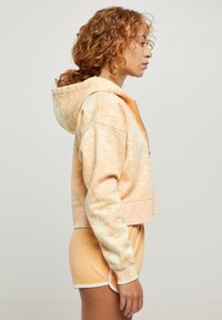 Hoodie cropped in pesca chiaro con motivo marmorizzato bianco, presenta un cappuccio, zip frontale e polsini a coste. Maniche corte visibili.