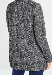 Abrigo texturizado en tonos gris y negro, con un corte holgado, cuello redondeado y detalles en la costura trasera. Hecho de una tela tejida con un patrón jaspeado.