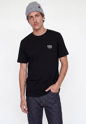 Jeune homme portant un bonnet gris, un t-shirt noir avec un logo blanc sur la poitrine, et un jean bleu foncé, debout devant un fond blanc.