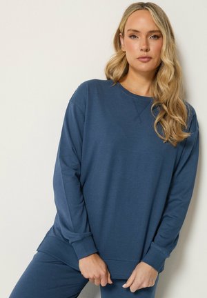 Kvinde med langt blondt hår iført en blå langærmet sweatshirt og matchende bukser, står mod en ensfarvet lys væg.