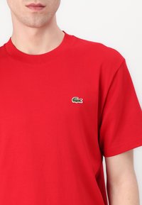 Homme portant un t-shirt rouge uni à manches courtes avec un petit logo crocodile vert sur la poitrine gauche.