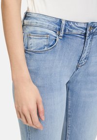 Jean en denim bleu clair avec une coupe slim, présentant un design traditionnel à cinq poches, des coutures visibles et une texture légèrement délavée.