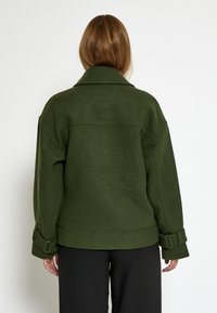 Žena s ravnom svijetlosmeškom kosom nosi tamnozeleni vuneni kaput s ovratnikom i detaljima kopčanja na rukavima, uparen s crnim hlačama.