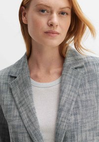 Gestructureerde grijze blazer met een notch lapel, gecombineerd met een lichtgrijze geribde shirt. De stof vertoont een visgraatpatroon. Schone, getailleerde silhouet.