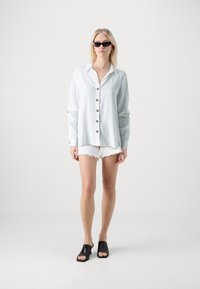 Helles blaues Button-Down-Hemd mit langen Ärmeln, kombiniert mit weißen, ausgefransten Shorts und schwarzen, offenen Mules. Model trägt schwarze Sonnenbrille.
