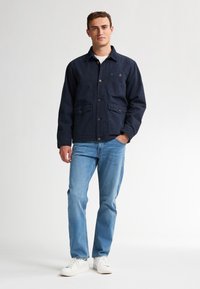Marineblauwe katoenen jack met patchzakken, knoopsluiting en een kraag, gedragen met lichtblauwe jeans en witte sneakers.