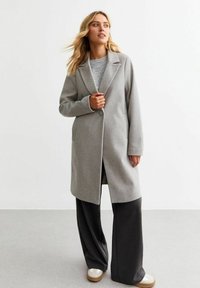 Grå ullblandad kappa med slagkrage, en knappstängning och två sidofickor, bärs över en grå tröja och svarta byxor.