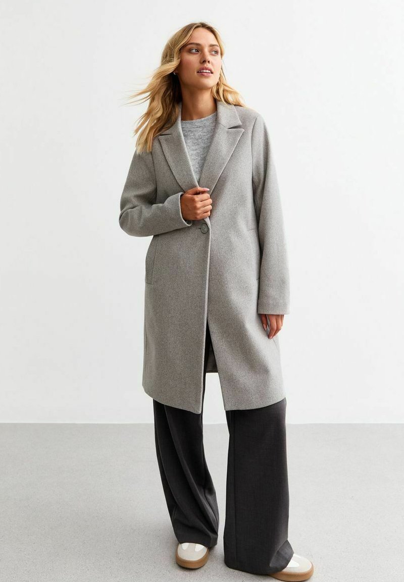 Grå ullblandad kappa med slagkrage, en knappstängning och två sidofickor, bärs över en grå tröja och svarta byxor.